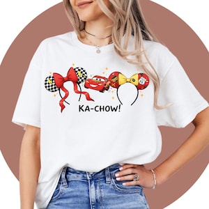 May include: White t-shirt featuring a design with a red bow, a red car, and a yellow bow with the text "KA-CHOW!". The design includes black and white checkered ears and a Route 66 sign. The shirt is a casual wear item.