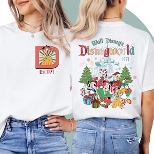 Könnte beinhalten: Weißes T-Shirt mit einem weihnachtlichen Design mit Mickey Mouse, Minnie Mouse, Goofy, Donald Duck und Pluto. Das Design enthält einen Weihnachtsbaum, Geschenke und den Text "Walt Disney's Disneyworld Est. 1971".
