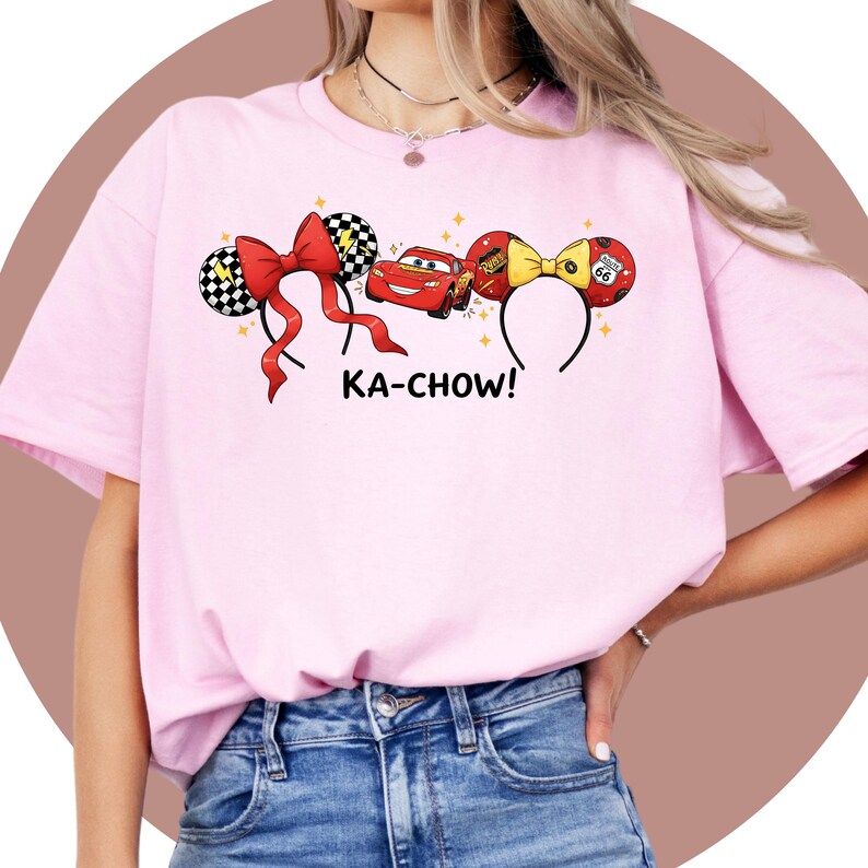 May include: Pink t-shirt featuring cartoon ears with red bows, a red race car, and the text "KA-CHOW!". The ears have black and white checkered patterns and Route 66 details. The shirt is a light pink color.