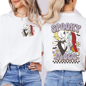Puede incluir: Camiseta blanca con un gr&aacute;fico de Jack Skellington y Sally de The Nightmare Before Christmas. La parte trasera de la camiseta tiene las palabras "Spooky Vibes" en una fuente retro, con una luna y detalles florales.