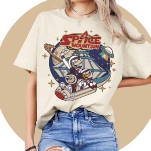 May include: A beige t-shirt featuring a vintage-style graphic of Mickey Mouse, Pluto, and Goofy riding a space mountain ride. The graphic includes the text "Space Mountain".