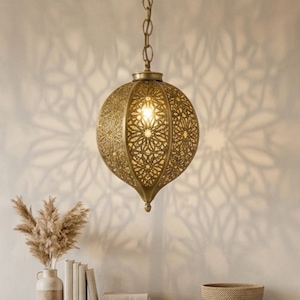 May include: A gold-colored, spherical pendant light with intricate cut-out patterns hangs from the ceiling, casting a decorative floral shadow on the wall. A wooden bench holds books, a vase with dried plants, and a woven bowl. A round jute rug sits on the floor.