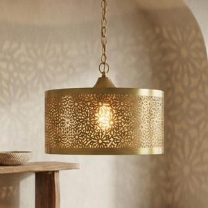 May include: A gold-colored, cylindrical pendant light fixture with intricate cut-out patterns. The light hangs from a chain and illuminates a warm glow. The fixture is suspended above a wooden console table and a woven rug.