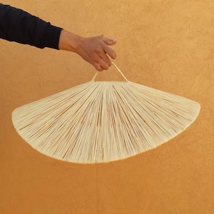 Può includere: Un grande paralume rotondo a forma di ventaglio, intrecciato in modo naturale, con un manico intrecciato. Il paralume è realizzato in fibre naturali di colore marrone chiaro ed è appeso contro un muro di colore arancio chiaro.