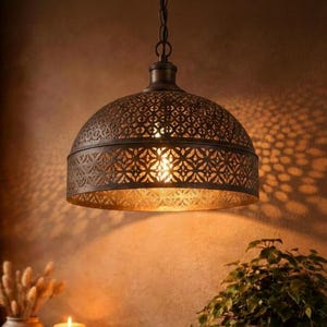 Op de afbeelding: Een bronzen hanglamp met een patroonkap hangt aan een ketting. Het licht werpt ingewikkelde schaduwen op de warm getinte muur. De afbeelding toont ook een dienblad met kaarsen, een vaas met gedroogde planten en een potplant.