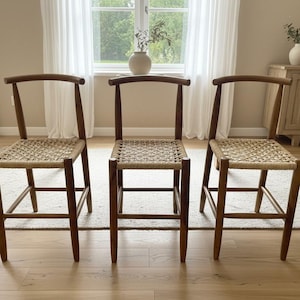 Könnte beinhalten: Drei Holzstühle mit gewebten Sitzen sind in einer Reihe angeordnet. Die Stühle haben eine geschwungene Rückenlehne und vier Beine. Die Sitze bestehen aus einem hellen, gewebten Material. Die Stühle stehen auf einem hellen Teppich.