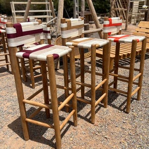 Op de afbeelding: Verschillende houten barkrukken met geweven zittingen en rugleuningen. De krukken hebben een natuurlijke houten afwerking en zijn versierd met rood, wit en beige geweven materiaal. De krukken staan buiten op een grindoppervlak.