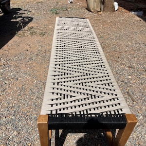May include: A long, rectangular bench with a woven seat in a black and beige pattern. The bench has a wooden frame and legs. The design is minimalist and suitable for indoor or outdoor use.