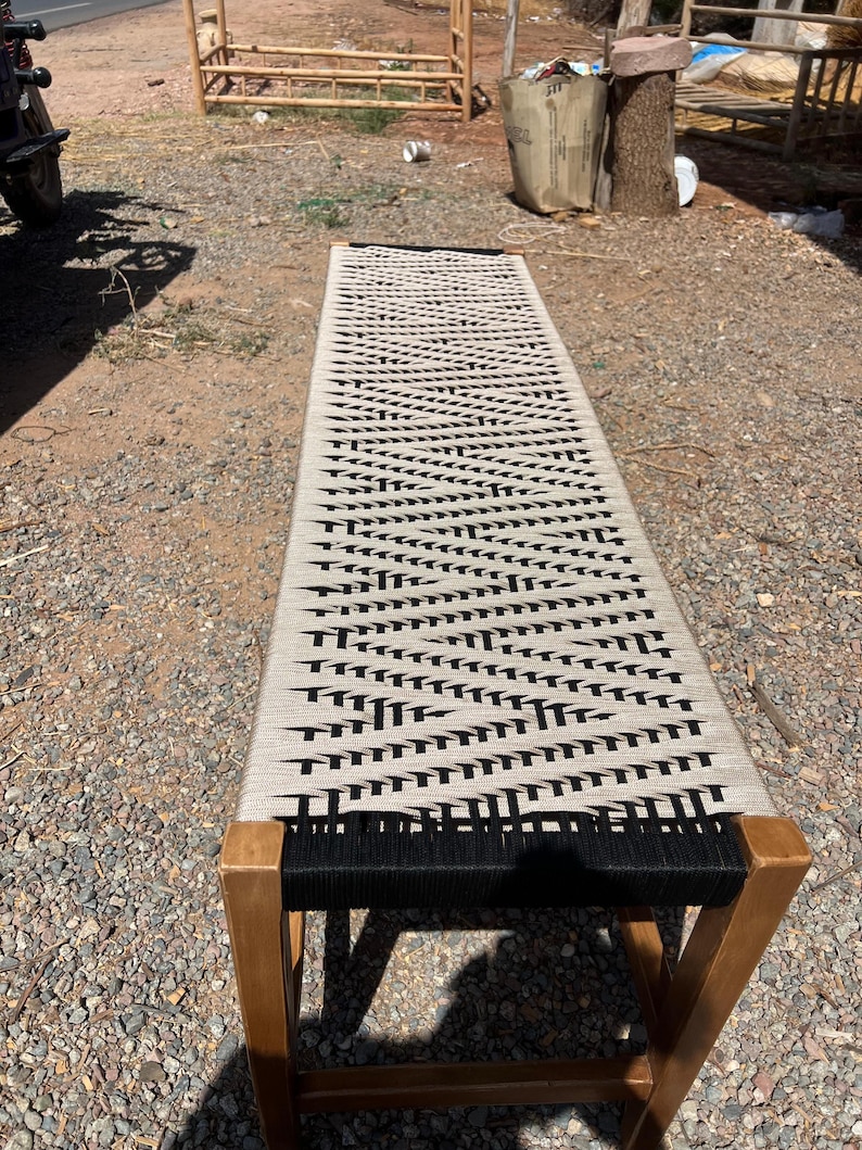 May include: A long, rectangular bench with a woven seat in a black and beige pattern. The bench has a wooden frame and legs. The design is minimalist and suitable for indoor or outdoor use.