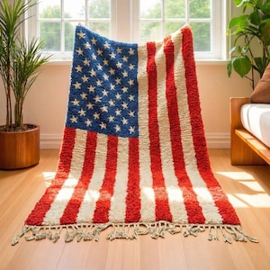 Puede incluir: Una alfombra con la forma de la bandera estadounidense, con rayas rojas y blancas y un campo azul con estrellas blancas. La alfombra tiene un aspecto texturizado y peludo y una franja a lo largo del borde inferior. Se muestra sobre un suelo de madera.