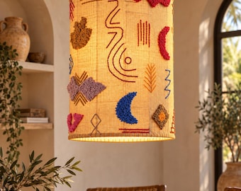 Puede incluir: Una pantalla de lámpara cilíndrica de tela color canela claro con bordados coloridos en relieve en rojo, azul, morado y amarillo mostaza. El diseño incluye formas abstractas, líneas y motivos de media luna cosidos en la superficie.