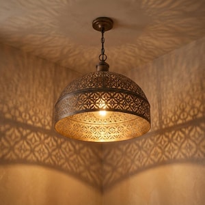 Pode incluir: Uma luminária pendente de cor bronze com uma cúpula estampada. A luz projeta sombras intrincadas nas paredes. A luminária pende de uma corrente e é iluminada por uma única lâmpada.