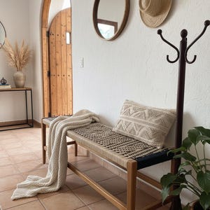 May include: A wooden bench with a woven seat and a cream-coloured throw blanket. A decorative cushion with a geometric pattern rests on the bench. A straw hat and oval mirror hang on the white wall.