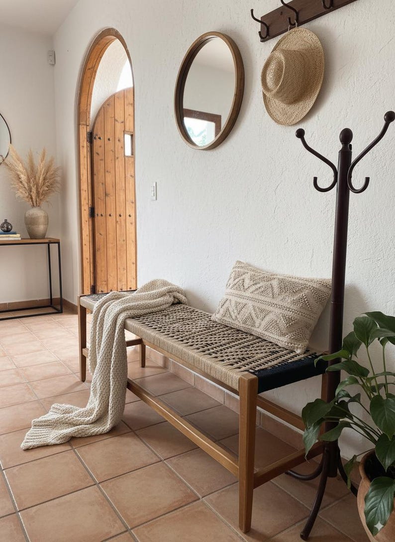 May include: A wooden bench with a woven seat and a cream-coloured throw blanket. A decorative cushion with a geometric pattern rests on the bench. A straw hat and oval mirror hang on the white wall.