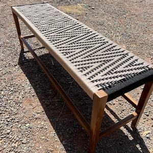 May include: A long wooden bench with a woven seat. The seat is made of a beige and black rope pattern. The bench has a rectangular shape and is supported by four wooden legs. The bench is outdoors.