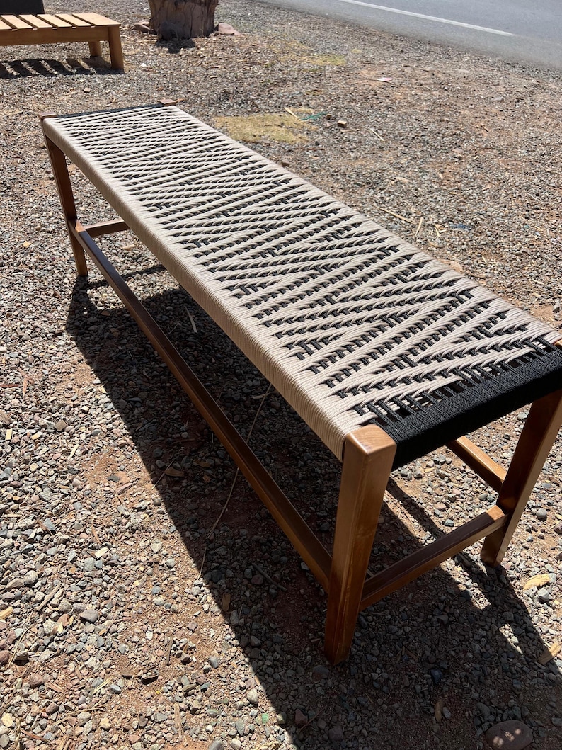 May include: A long wooden bench with a woven seat. The seat is made of a beige and black rope pattern. The bench has a rectangular shape and is supported by four wooden legs. The bench is outdoors.