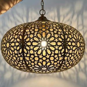 May include: A brass-colored, oval-shaped pendant light fixture with intricate star and geometric cut-out patterns. The light shines through the cutouts, casting star-shaped shadows. The fixture hangs from a chain.