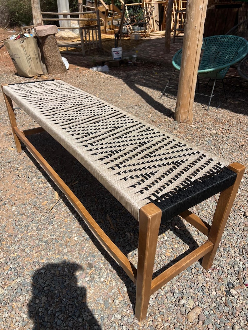 May include: A long wooden bench with a woven seat. The seat is made of beige and black rope, and the frame is made of light brown wood. The bench is outdoors on a gravel surface.