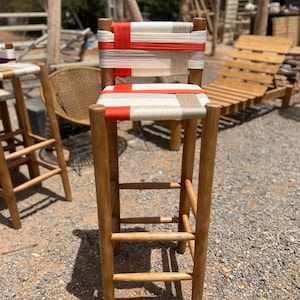 Könnte beinhalten: Ein hoher Barhocker aus Holz mit gewebter Sitzfläche und Rückenlehne. Sitzfläche und Rückenlehne weisen ein Muster aus weißem, beigem und rotem gewebtem Material auf. Der Hocker hat vier Holzbeine und Fußstützen. Im Hintergrund sind weitere Holzmöbel zu sehen.