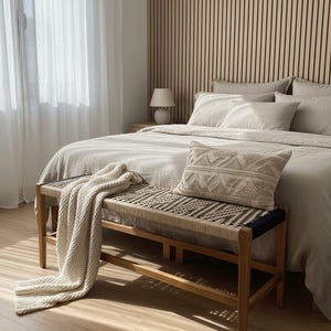 May include: A bedroom interior with a bed dressed in neutral-toned bedding, a woven bench, and a decorative pillow. A cream-coloured throw blanket is draped on the bench. The room features a wooden accent wall and sheer curtains.