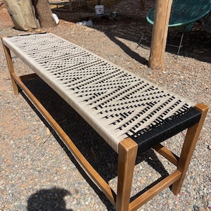May include: A long wooden bench with a woven seat. The seat is made of beige and black rope, with a repeating pattern. The legs and frame are made of light brown wood. The bench is outdoors.