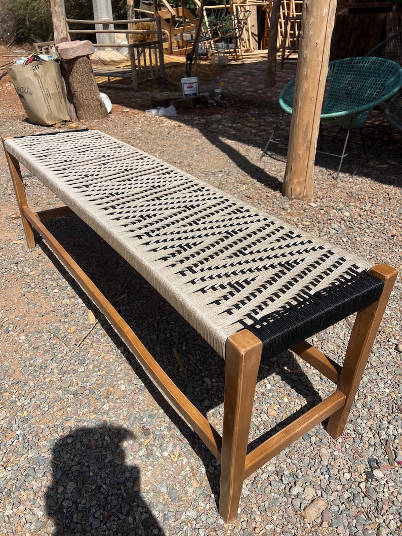May include: A long wooden bench with a woven seat. The seat is made of beige and black rope, with a repeating pattern. The legs and frame are made of light brown wood. The bench is outdoors.