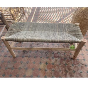 May include: A wooden bench with a woven seat made of natural fibers. The bench has a simple design with four legs and a rectangular seat. The bench is sitting on a tiled floor.