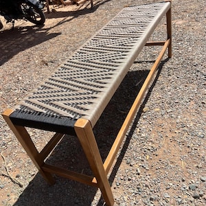 May include: A long wooden bench with a woven seat. The seat is made of alternating black and beige rope. The bench has a rectangular shape and is supported by wooden legs. The bench is outdoors on a gravel surface.