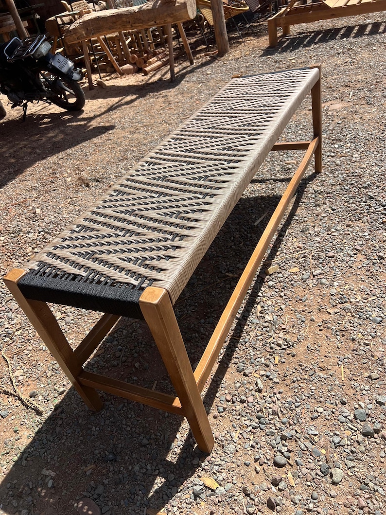 May include: A long wooden bench with a woven seat. The seat is made of alternating black and beige rope. The bench has a rectangular shape and is supported by wooden legs. The bench is outdoors on a gravel surface.