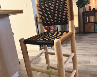 May include: A wooden bar stool with a woven black and brown seat and back. The stool has a natural wood finish and a simple design.