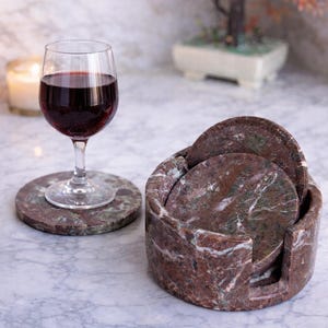 May include: A set of marble coasters and a holder. The coasters are round, with red, brown, and green veining. A wine glass with red wine sits on one coaster. The holder is cylindrical. Coasters are approximately 10 cm in diameter.