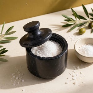 May include: A cylindrical black marble salt cellar with a matching lid, filled with coarse white sea salt. Beside it sits a small, round, off-white ceramic bowl also containing white sea salt, accompanied by green olive branches.