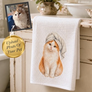 May include: White waffle-weave towel featuring a watercolor illustration of a ginger and white cat wearing a gray towel on its head. The towel is hanging on a distressed white cabinet. A framed photo of a cat and text "Upload Photo Of Your Pet" are also visible.