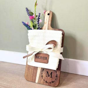 May include: A wooden cutting board with a handle, featuring a light-colored stripe down the middle. A white towel, wooden spoon, and a cream-colored ribbon are tied around the board. The board has the name "MOYER" and the letter "M" engraved on it. Flowers are placed in the handle.