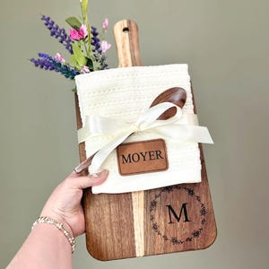 May include: A wooden cutting board with a handle, a white towel, and a wooden spoon tied with a cream ribbon. The board has a floral wreath with the letter "M" and a leather patch that says "MOYER". Flowers are placed in the handle.