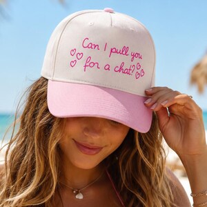 May include: A baseball cap with a cream-colored crown and a pink brim. The front of the cap features the embroidered pink text "Can I pull you for a chat?" surrounded by small embroidered pink heart icons on both sides of the text.