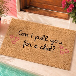 May include: A rectangular tan coir doormat featuring the black text Can I pull you for a chat? written in a cursive script. The mat is decorated with five small pink heart outlines scattered around the text on the textured surface.