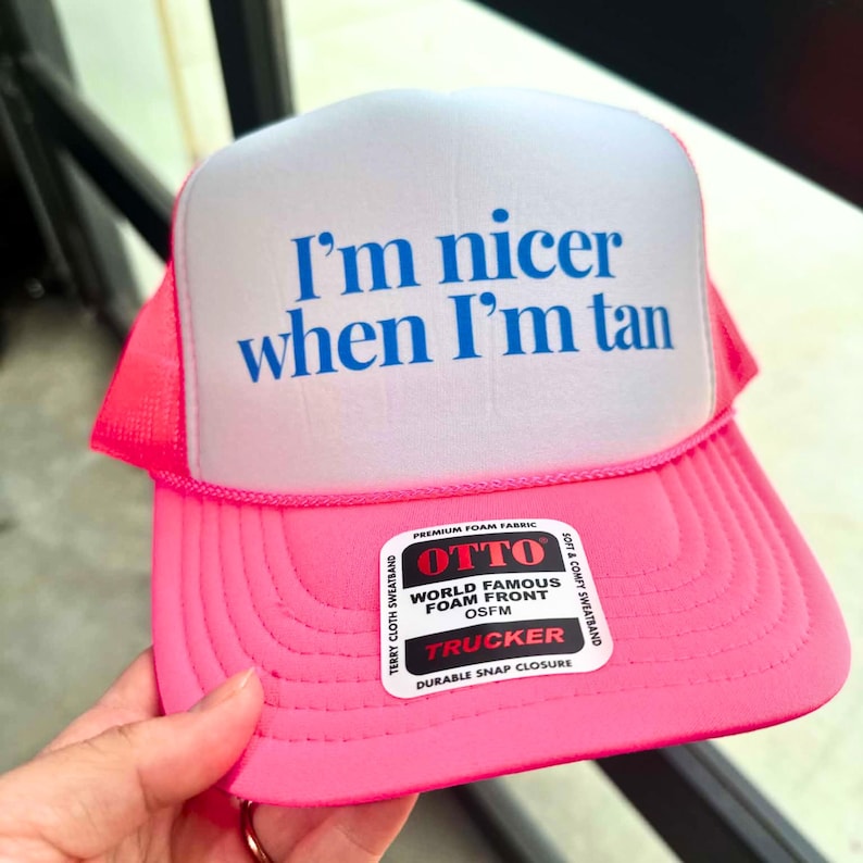May include: A bright pink and white trucker hat featuring a foam front panel with the blue text I'm nicer when I'm tan. The hat includes a pink mesh back, a pink brim with stitching, and a white sticker label with black text reading OTTO.