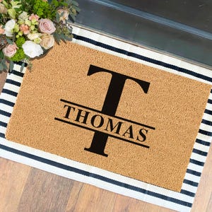 May include: A brown coir doormat with black stripes around the edges. The doormat has a large black letter "T" with the name "THOMAS" written in black letters below it.