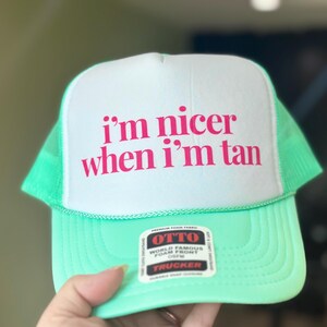 May include: A bright mint green trucker hat with a white foam front panel. The text "i'm nicer when i'm tan" is printed in bold pink letters on the front. An Otto brand sticker is attached to the brim, featuring black, white, and red text.