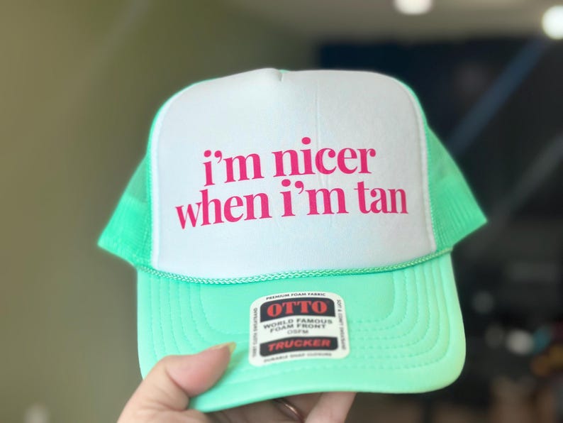 May include: A bright mint green trucker hat with a white foam front panel. The text "i'm nicer when i'm tan" is printed in bold pink letters on the front. An Otto brand sticker is attached to the brim, featuring black, white, and red text.