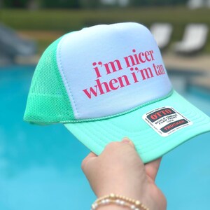 May include: A mint green and white trucker hat featuring a white foam front panel with the text i'm nicer when i'm tan printed in pink. The hat includes a mesh back, a solid mint green brim, and a black and white sticker on the brim.