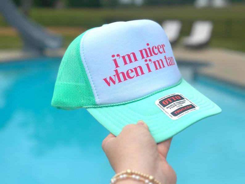May include: A mint green and white trucker hat featuring a white foam front panel with the text i'm nicer when i'm tan printed in pink. The hat includes a mesh back, a solid mint green brim, and a black and white sticker on the brim.