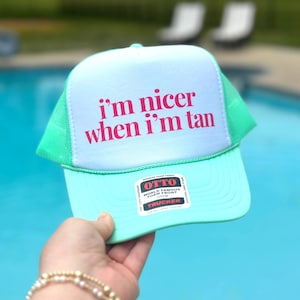 May include: A mint green and white trucker hat featuring the text i'm nicer when i'm tan in bold pink lettering on the front foam panel. The cap includes a mesh back, a curved brim, and a rectangular product sticker on the lower front section.