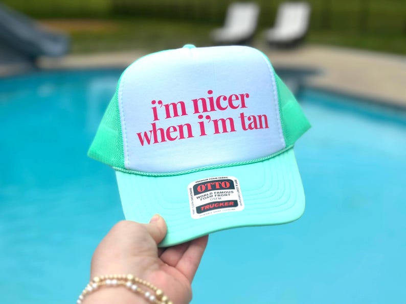 May include: A mint green and white trucker hat featuring the text i'm nicer when i'm tan in bold pink lettering on the front foam panel. The cap includes a mesh back, a curved brim, and a rectangular product sticker on the lower front section.