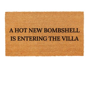 May include: A rectangular doormat made of natural brown coir fibre. The mat features the text "A HOT NEW BOMBSHELL IS ENTERING THE VILLA" in black, centered on the mat. The background is plain white.