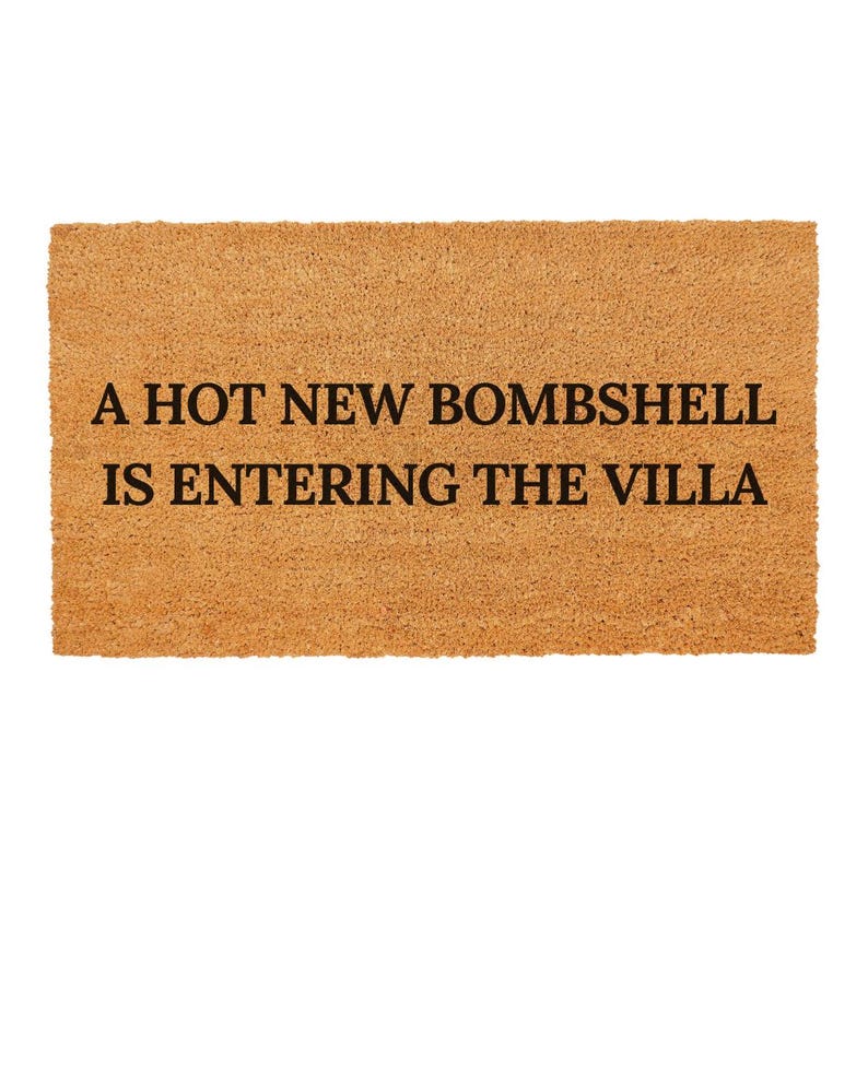 May include: A rectangular doormat made of natural brown coir fibre. The mat features the text "A HOT NEW BOMBSHELL IS ENTERING THE VILLA" in black, centered on the mat. The background is plain white.