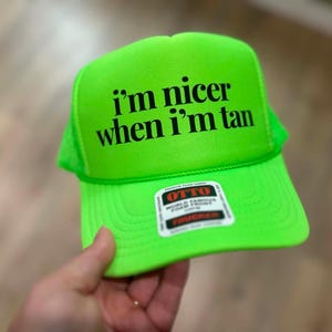 May include: A neon green trucker hat featuring the black text i'm nicer when i'm tan printed on the front foam panel. The cap includes a structured front, a curved brim, and a small rectangular sticker with the brand name OTTO in red and black.