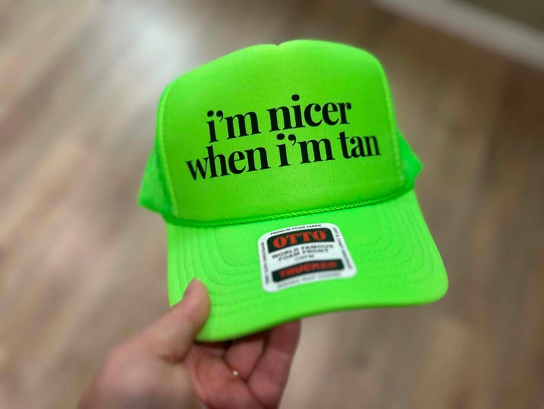 May include: A neon green trucker hat featuring the black text i'm nicer when i'm tan printed on the front foam panel. The cap includes a structured front, a curved brim, and a small rectangular sticker with the brand name OTTO in red and black.