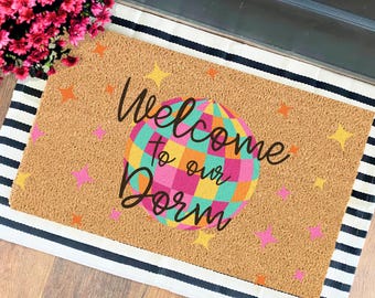 Op de afbeelding: Een bruine kokosvezel deurmat met zwarte en witte strepen rond de randen. De deurmat heeft een kleurrijk discobal ontwerp met de tekst "Welcome to our Dorm" in zwart.